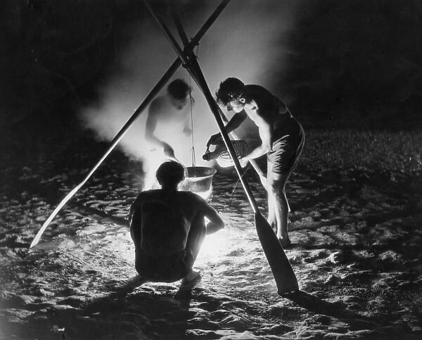 CAMP FIRE. Three young men cooking a meal on a beach