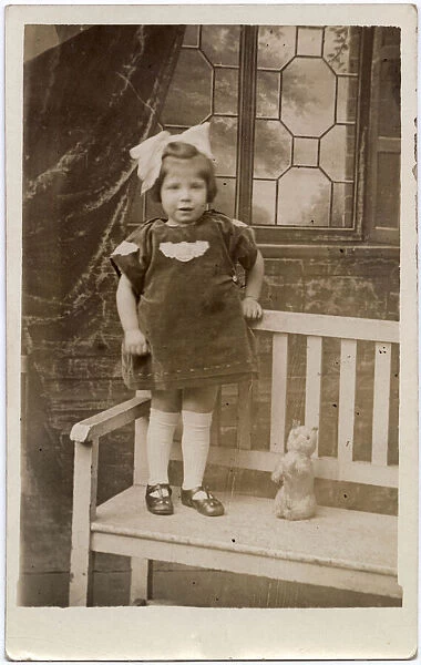 A little girl with an enormous bow in her hair stands on a garden bench with her toy cat