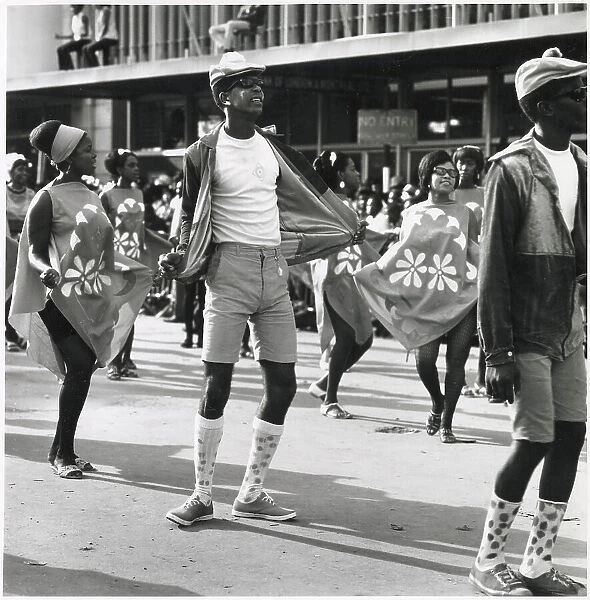 A man in shorts and long socks dances between two fashionable women at the Port of Spain Carnival, Trinidad, West Indies. Date: 1968