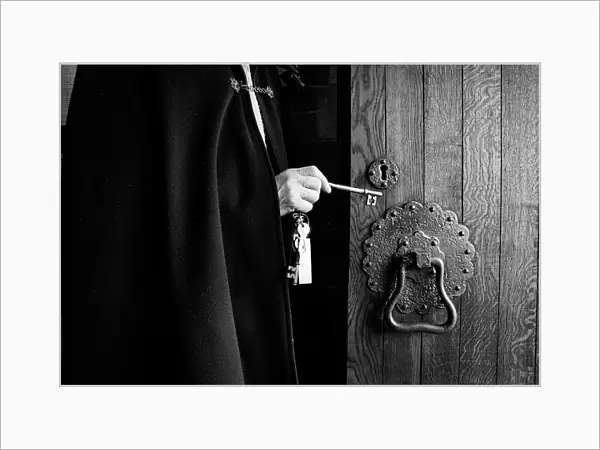A priest wearing a black cloak unlocks the oak door of a chu