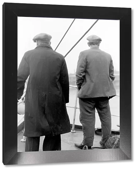 Life on a North Sea trawler - two men on deck in raincoat, jacket, and flat caps, close to shore. Date: 1960s