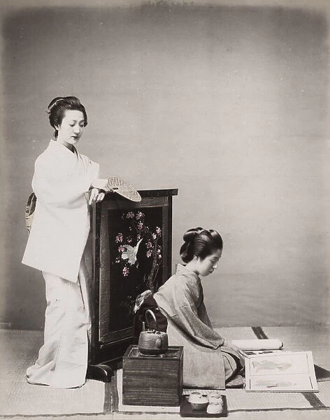 Two geishas, one reading, with stove a tea set. Japan