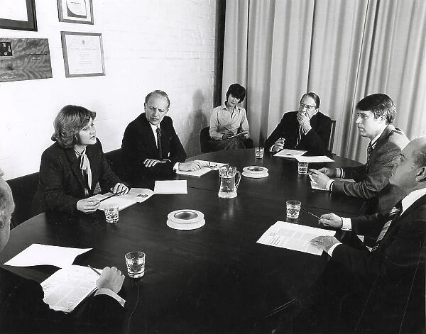 A group of men and women sit around a table holding a board meeting. A (female) secretary takes notes. Date: circa 1970