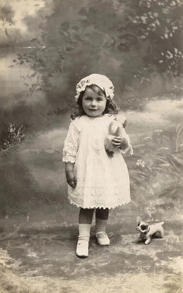 A little girl in lacy dress and cap poses with a toy bulldog Date: circa 1915