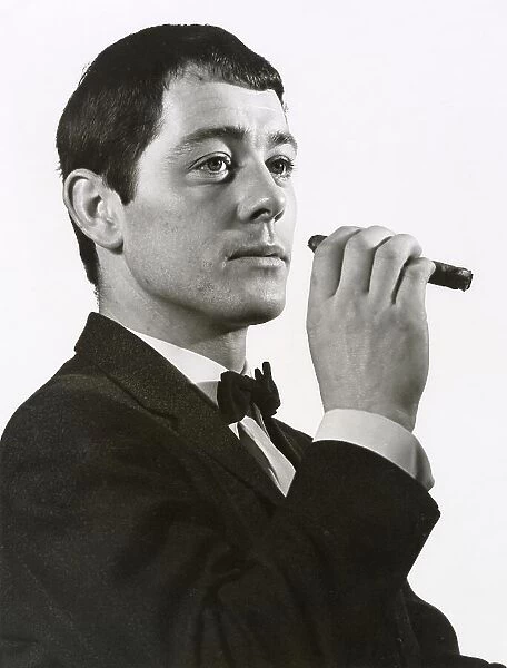 A man poses in a dinner jacket smoking a cigar. Date: late 1960s