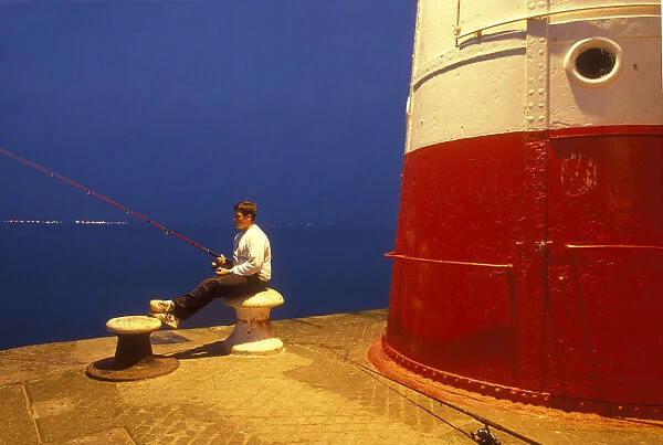 Teenage boy sits on a bollard while fishing, Newlyn Harbour