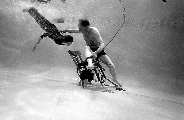 Underwater escapologist - a boy dives into a swimming pool to help a man untie himself from a chair. (1 of 3) Date: 1967