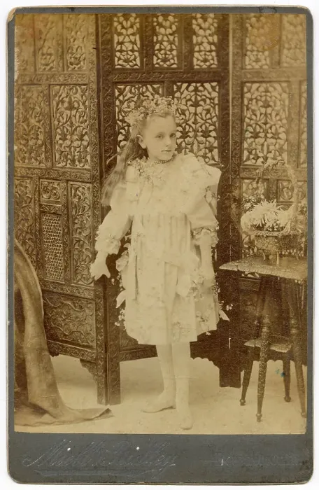 Framed Print : Young girl in studio photo with ornately carved wooden screen behind her Date: circa 1910s