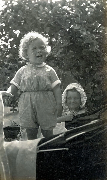 Two young siblings, one standing in a fine baby carriage