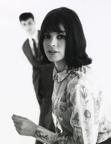 A young woman poses in the foreground with an out of focus male standing behind. Date: mid 1960s