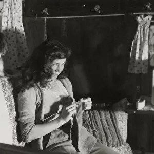 A mischievous-looking girl, caught on camera knitting in an armchair in her cottage