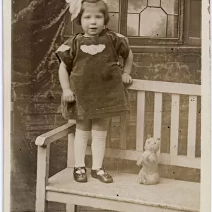 TOY CAT, a little girl with an enormous bow in her hair stands on a garden bench with her