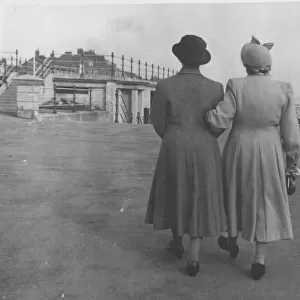 Back view of two ladies walking along an unidentified sea front arm in arm. Date: c