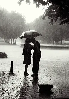 Atmospheric shot of young female-male couple under umbrella in rain in park, heads inclined to each other; has romantic feel Date: early 1950s
