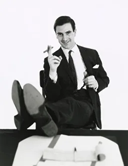 A man puts his feet up on his desk as he smokes a cigar. Date: late 1960s