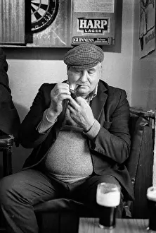 Guinness Prints: Mature man wearing cloth cap lights his pipe in an Irish pub